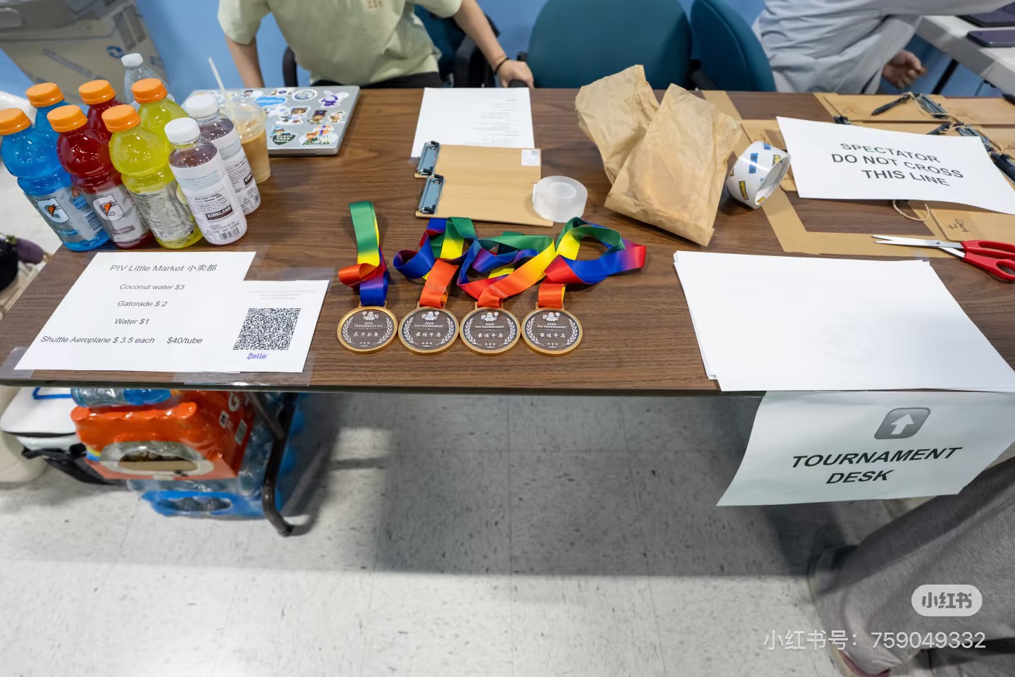 Medals with rainbow ribbons laid out on the tournament desk beside drinks and the PIV Little Market price list