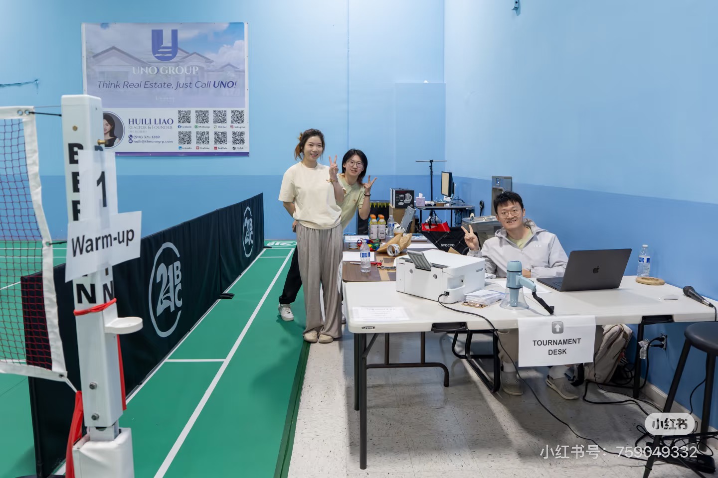 PIV volunteers waving from the tournament desk, with laptop, printer, and megaphone set up next to the warm-up court